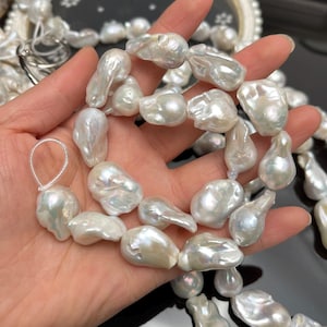 May include: A strand of large, irregularly shaped white baroque pearls. The pearls have a lustrous, iridescent sheen and vary in size and form. The pearls are strung together, creating a necklace or bracelet. The background is dark, highlighting the pearls.