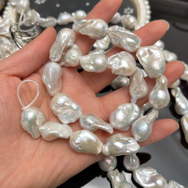 May include: A strand of large, irregularly shaped white baroque pearls. The pearls have a lustrous, iridescent sheen and vary in size and form. The pearls are strung together, creating a necklace or bracelet. The background is dark, highlighting the pearls.