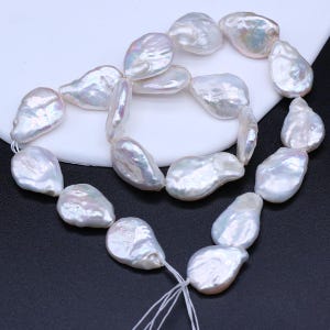 Puede incluir: Un collar de perlas barrocas de forma irregular. Las perlas son blancas con sutiles tonos rosas y azules, ensartadas en un hilo blanco. Las formas únicas de las perlas les dan un aspecto orgánico. Las perlas se muestran sobre una superficie blanca y negra.