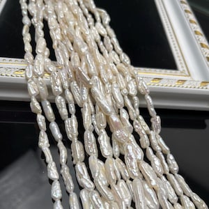 May include: A close-up of multiple strands of white, oblong-shaped pearls. The pearls have a lustrous, iridescent sheen and are arranged in a cascading pattern. The background includes a white and gold-trimmed frame.