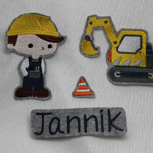 Könnte beinhalten: Ein graues Filznamenschild mit dem Namen "Jannik", der in blauer Farbe gestickt ist. Es gibt drei gestickte Aufnäher: ein gelber Bauarbeiter mit einem braunen Werkzeuggürtel, ein gelber Bagger und ein orangefarbener Verkehrskegel.