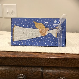 May include: A hand-painted wooden angel ornament with a white gown, gold halo, and brown hair. The angel is flying against a blue background with white stars and snowflakes.