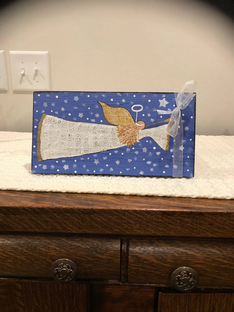May include: A hand-painted wooden angel ornament with a white gown, gold halo, and brown hair. The angel is flying against a blue background with white stars and snowflakes.