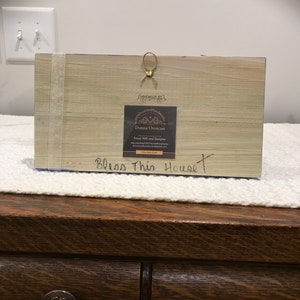 May include: A wooden sign with the text "Bless This House" painted in black. The sign is decorated with a blue and white floral design. A small, gold-colored tag with the text "Old American Handmades" and the name "Donna Overcast" is attached to the sign.