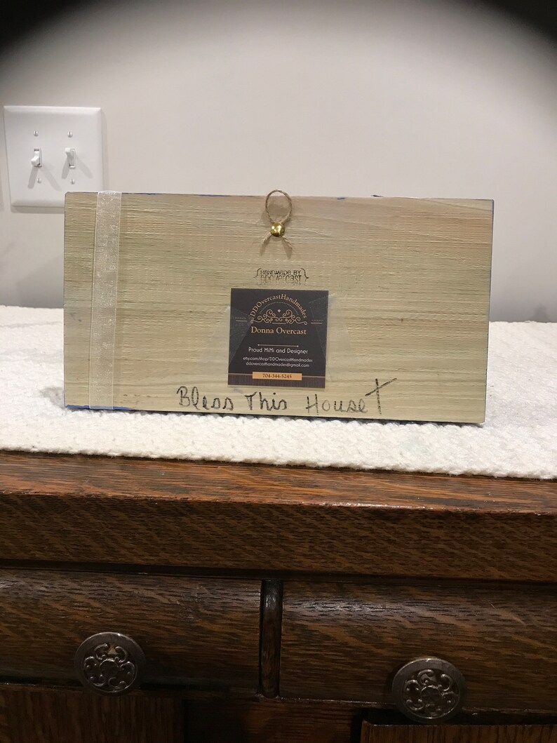 May include: A wooden sign with the text "Bless This House" painted in black. The sign is decorated with a blue and white floral design. A small, gold-colored tag with the text "Old American Handmades" and the name "Donna Overcast" is attached to the sign.