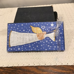 May include: A hand-painted wooden sign featuring a white angel with gold wings and a halo, blowing a trumpet. The angel is set against a blue background with white stars and snowflakes.