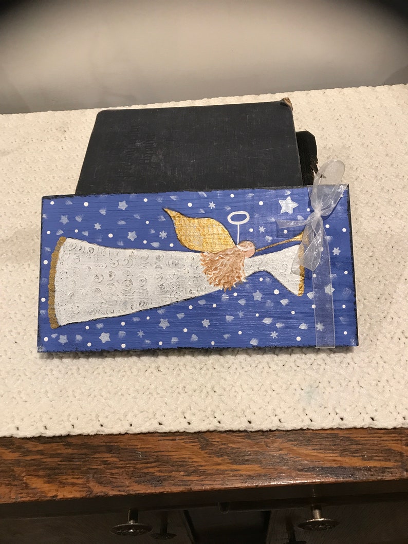May include: A hand-painted wooden sign featuring a white angel with gold wings and a halo, blowing a trumpet. The angel is set against a blue background with white stars and snowflakes.