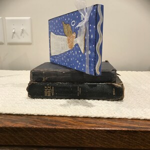 May include: A wooden angel figurine painted on a blue background with white stars and a white ribbon. The angel has a halo and is holding a white banner. The figurine is sitting on top of two black leather-bound Bibles.