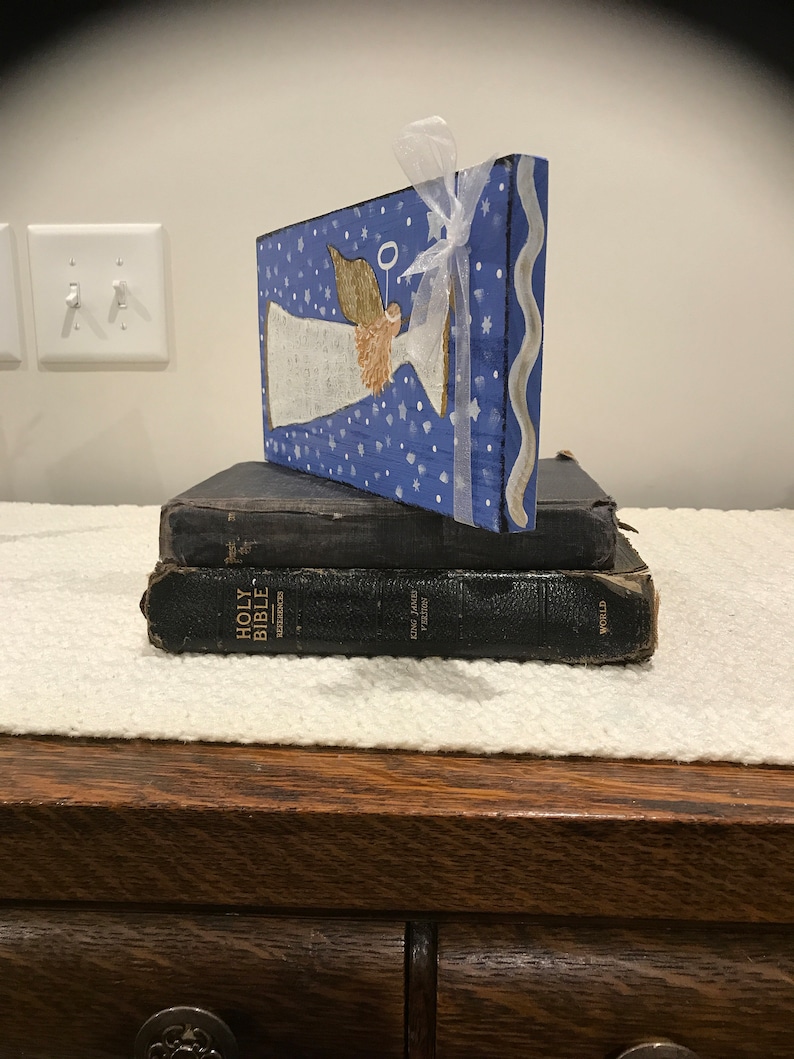 May include: A wooden angel figurine painted on a blue background with white stars and a white ribbon. The angel has a halo and is holding a white banner. The figurine is sitting on top of two black leather-bound Bibles.