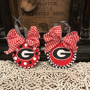 May include: Two red, white, and black painted wooden ornaments with a white and red gingham bow. Each ornament features a white and black polka dot background with a black and red "G" in the center. The ornaments are sitting on a white crocheted doily.
