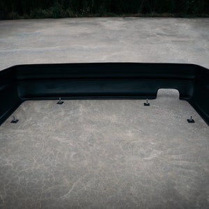 May include: Black plastic rear bumper for a car. The bumper is sitting on a concrete surface and has several mounting points.