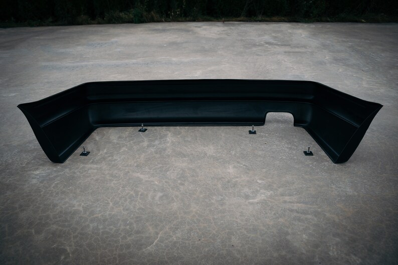 May include: Black plastic rear bumper for a car. The bumper is sitting on a concrete surface and has several mounting points.