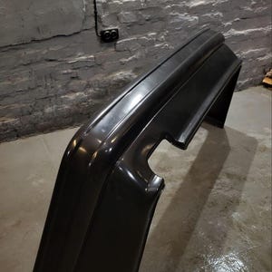 Puede incluir: Un parachoques negro brillante para autom&oacute;vil, probablemente para la parte trasera de un veh&iacute;culo, est&aacute; inclinado contra una pared de ladrillos texturizada y de color claro. El parachoques tiene un dise&ntilde;o curvo con una secci&oacute;n recortada.