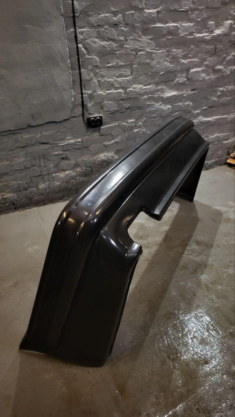 Puede incluir: Un parachoques negro brillante para autom&oacute;vil, probablemente para la parte trasera de un veh&iacute;culo, est&aacute; inclinado contra una pared de ladrillos texturizada y de color claro. El parachoques tiene un dise&ntilde;o curvo con una secci&oacute;n recortada.