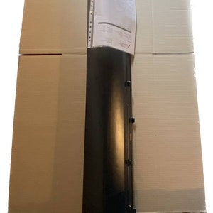 K&ouml;nnte beinhalten: Ein langes, schwarzes, rechteckiges Objekt mit einer Reihe kleiner, dunkler Befestigungselemente an einer Seite. Ein wei&szlig;es Papier mit Text ist in der Mitte befestigt. Das Objekt liegt auf einem Stapel Pappkartons.