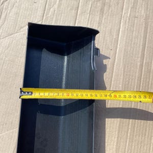 K&ouml;nnte beinhalten: Ein schwarzes Kunststoff-Autoteil, m&ouml;glicherweise ein Zierteil, wird mit einem gelben Ma&szlig;band gezeigt. Das Band zeigt eine L&auml;nge von etwa 25 cm an. Das Teil hat eine rechteckige Form mit einer gebogenen Kante und einer strukturierten Oberfl&auml;che.