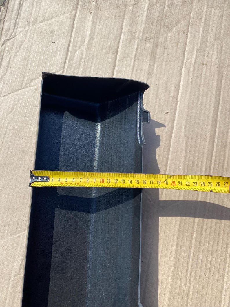 K&ouml;nnte beinhalten: Ein schwarzes Kunststoff-Autoteil, m&ouml;glicherweise ein Zierteil, wird mit einem gelben Ma&szlig;band gezeigt. Das Band zeigt eine L&auml;nge von etwa 25 cm an. Das Teil hat eine rechteckige Form mit einer gebogenen Kante und einer strukturierten Oberfl&auml;che.