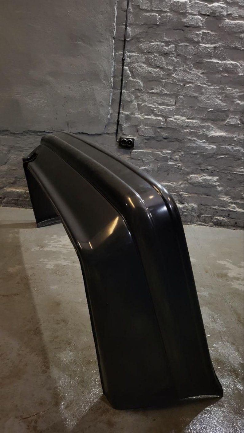 Puede incluir: Un parachoques negro brillante con un dise&ntilde;o curvo. El parachoques est&aacute; en &aacute;ngulo, mostrando su forma y superficie reflectante. El fondo presenta una pared texturizada de color claro y un suelo de hormig&oacute;n.