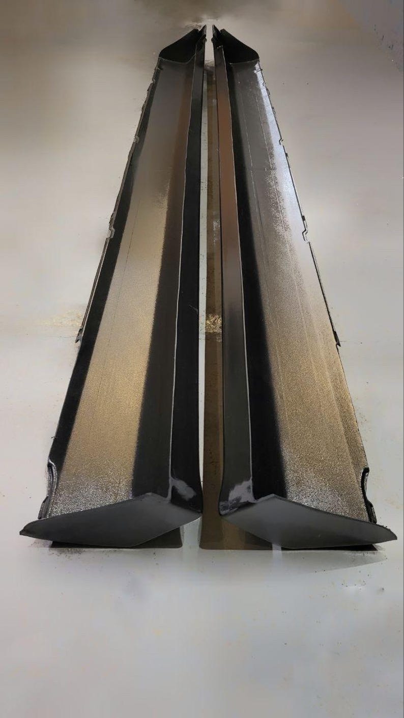 K&ouml;nnte beinhalten: Zwei schwarze, kantige Seitenschweller f&uuml;r Automobile, vertikal positioniert und zueinander geneigt. Die Oberfl&auml;chen haben eine gl&auml;nzende Oberfl&auml;che, die das Licht reflektiert. Das Design deutet auf einen Fokus auf Fahrzeug&auml;sthetik und Aerodynamik hin.