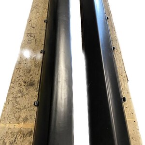 K&ouml;nnte beinhalten: Zwei schwarze Seitenschweller f&uuml;r Autos, wahrscheinlich f&uuml;r die Schweller eines Fahrzeugs, werden gezeigt. Sie sind lang, rechteckig und haben ein leicht abgewinkeltes Design. Die Teile liegen auf einer Holzoberfl&auml;che und sind bereit f&uuml;r die Installation.