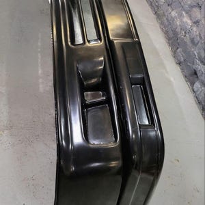 Puede incluir: Se muestran dos parachoques de coche negros, con un acabado brillante. Los parachoques tienen forma rectangular con aberturas y contornos integrados, lo que sugiere un dise&ntilde;o automotriz moderno. La imagen est&aacute; tomada desde un &aacute;ngulo ligeramente elevado.