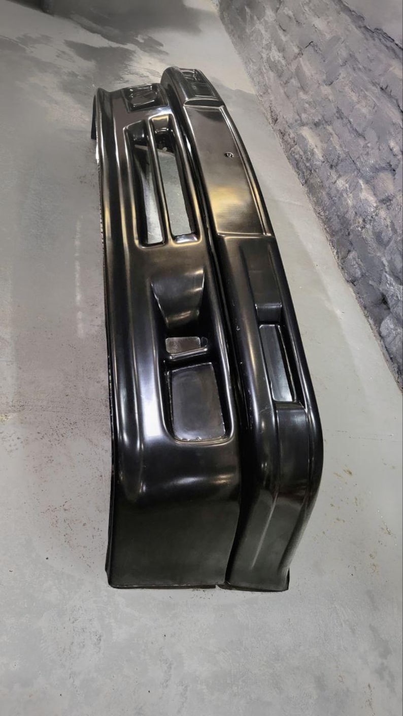 Puede incluir: Se muestran dos parachoques de coche negros, con un acabado brillante. Los parachoques tienen forma rectangular con aberturas y contornos integrados, lo que sugiere un dise&ntilde;o automotriz moderno. La imagen est&aacute; tomada desde un &aacute;ngulo ligeramente elevado.