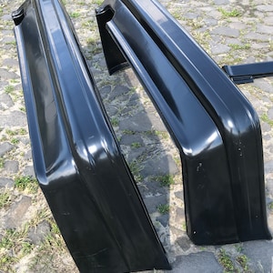 May include: Two black plastic car bumpers, one is lying on its side and the other is standing upright. The bumpers are made of a smooth, glossy material.