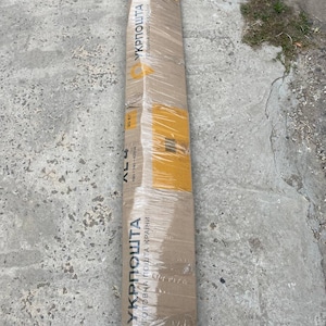 K&ouml;nnte beinhalten: Ein langes, rechteckiges Kartonpaket, das in transparente Folie verpackt ist. Das Paket hat gelbes Klebeband und den ukrainischen Text "УКРПОШТА". Es liegt auf einer grauen Betonoberfl&auml;che.