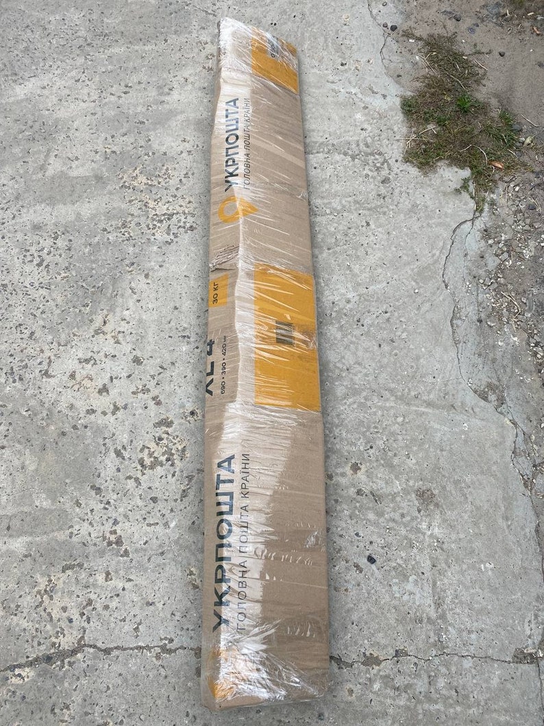 K&ouml;nnte beinhalten: Ein langes, rechteckiges Kartonpaket, das in transparente Folie verpackt ist. Das Paket hat gelbes Klebeband und den ukrainischen Text "УКРПОШТА". Es liegt auf einer grauen Betonoberfl&auml;che.
