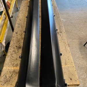 K&ouml;nnte beinhalten: Zwei schwarze Seitenschweller f&uuml;r Autos werden auf einer Holzoberfl&auml;che gezeigt. Die langen, rechteckigen Teile haben ein leicht abgewinkeltes Design und bestehen aus einem glatten, dunklen Material. Sie scheinen neu oder k&uuml;rzlich &uuml;berholt zu sein.