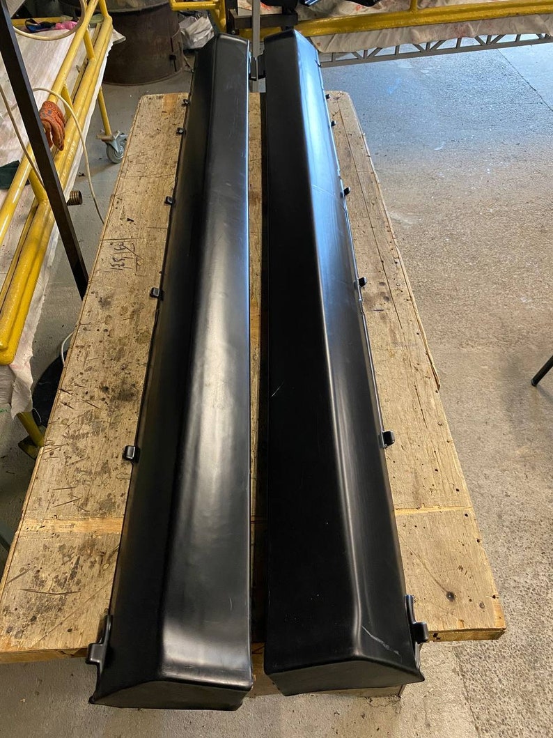 K&ouml;nnte beinhalten: Zwei schwarze Seitenschweller f&uuml;r Autos werden auf einer Holzoberfl&auml;che gezeigt. Die langen, rechteckigen Teile haben ein leicht abgewinkeltes Design und bestehen aus einem glatten, dunklen Material. Sie scheinen neu oder k&uuml;rzlich &uuml;berholt zu sein.