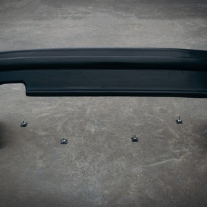 May include: Black plastic rear bumper for a car. The bumper is sitting on a gray concrete surface. There are five small black plastic fasteners on the surface.