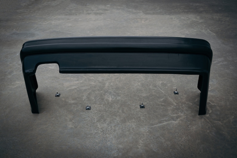May include: Black plastic rear bumper for a car. The bumper is sitting on a gray concrete surface. There are five small black plastic fasteners on the surface.