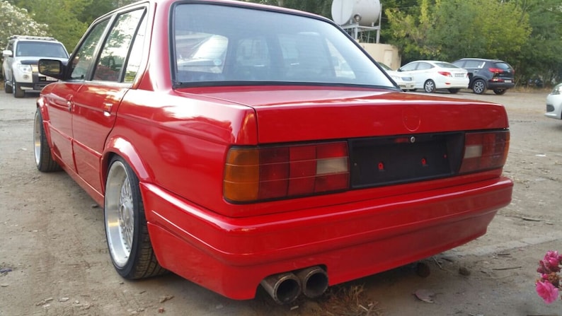 May include: A red BMW 3 Series sedan with a black rear bumper and a black license plate. The car has a chrome exhaust tip and a silver wheel.