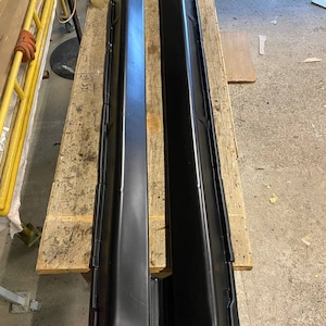 K&ouml;nnte beinhalten: Zwei schwarze Seitenschweller f&uuml;r Autos werden auf einer Holzpalette pr&auml;sentiert. Die Seitenschweller sind lang, rechteckig und haben ein leicht abgewinkeltes Design. Sie bestehen aus einem gl&auml;nzenden, dunklen Material und scheinen neu zu sein.