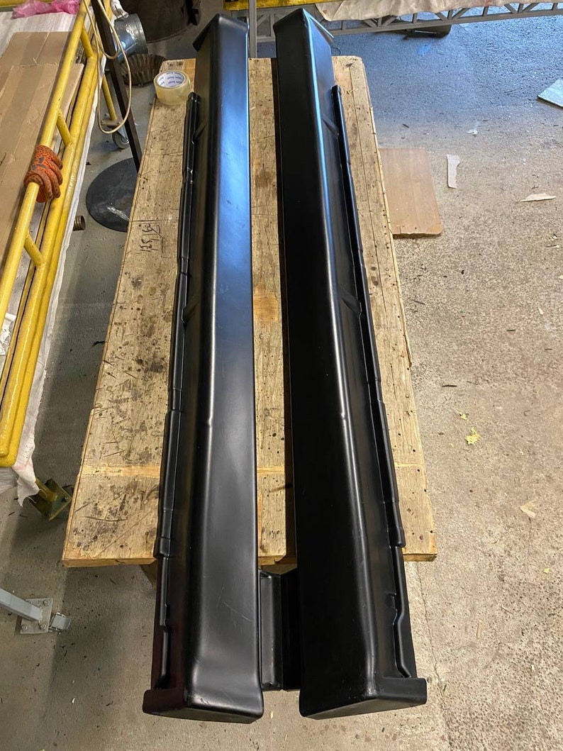 K&ouml;nnte beinhalten: Zwei schwarze Seitenschweller f&uuml;r Autos werden auf einer Holzpalette pr&auml;sentiert. Die Seitenschweller sind lang, rechteckig und haben ein leicht abgewinkeltes Design. Sie bestehen aus gl&auml;nzendem schwarzem Material und scheinen neu zu sein.