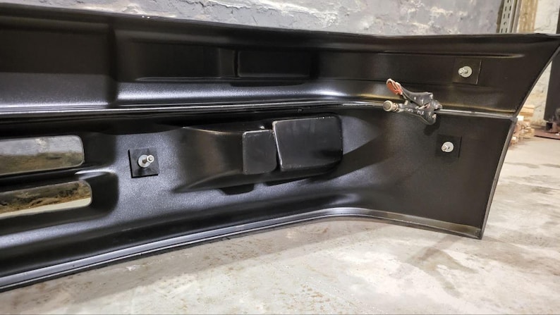 Puede incluir: Un parachoques automotriz negro con un dise&ntilde;o curvo. El parachoques tiene varios recortes rectangulares y est&aacute; asegurado con pernos y una abrazadera. La superficie tiene un acabado brillante, que refleja el entorno.