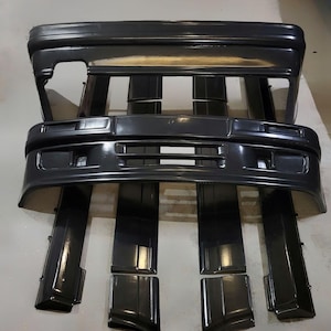 Puede incluir: Un conjunto de parachoques y molduras de coche negros. Los parachoques tienen forma rectangular con aberturas para luces y una rejilla. Las molduras son largas y estrechas, dise&ntilde;adas para encajar a lo largo de los lados del coche. Las piezas est&aacute;n dispuestas sobre una superficie gris claro.