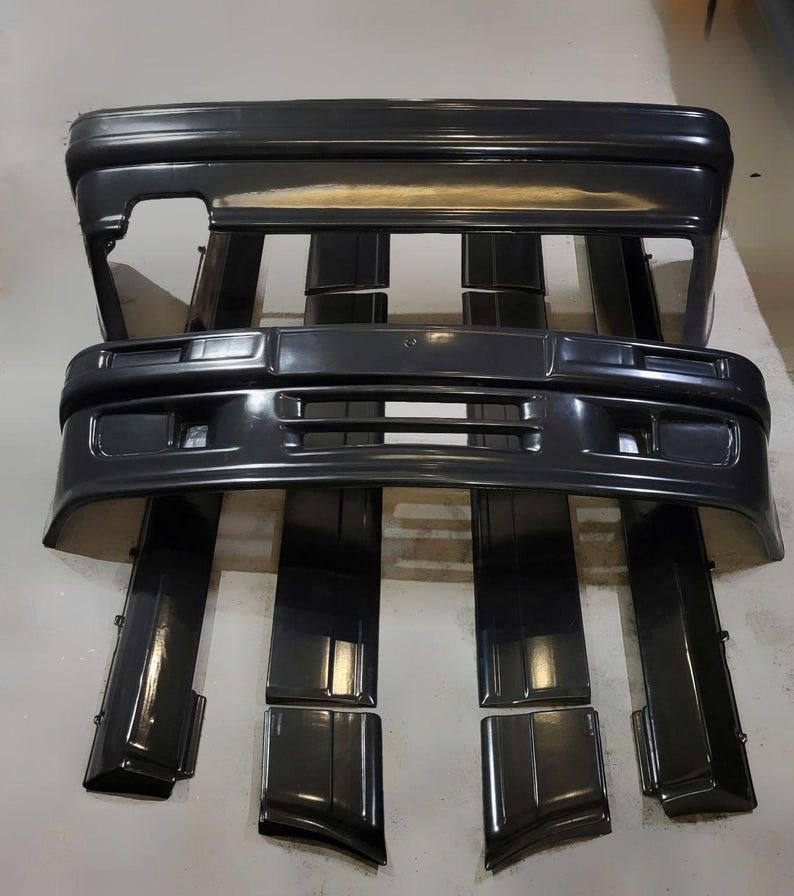 Puede incluir: Un conjunto de parachoques y molduras de coche negros. Los parachoques tienen forma rectangular con aberturas para luces y una rejilla. Las molduras son largas y estrechas, dise&ntilde;adas para encajar a lo largo de los lados del coche. Las piezas est&aacute;n dispuestas sobre una superficie gris claro.