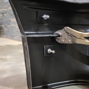 Puede incluir: Primer plano de una secci&oacute;n de parachoques automotriz negra con sujetadores met&aacute;licos. Un par de alicates est&aacute; sujeto al borde del parachoques. El parachoques est&aacute; contra una pared de ladrillos gris.