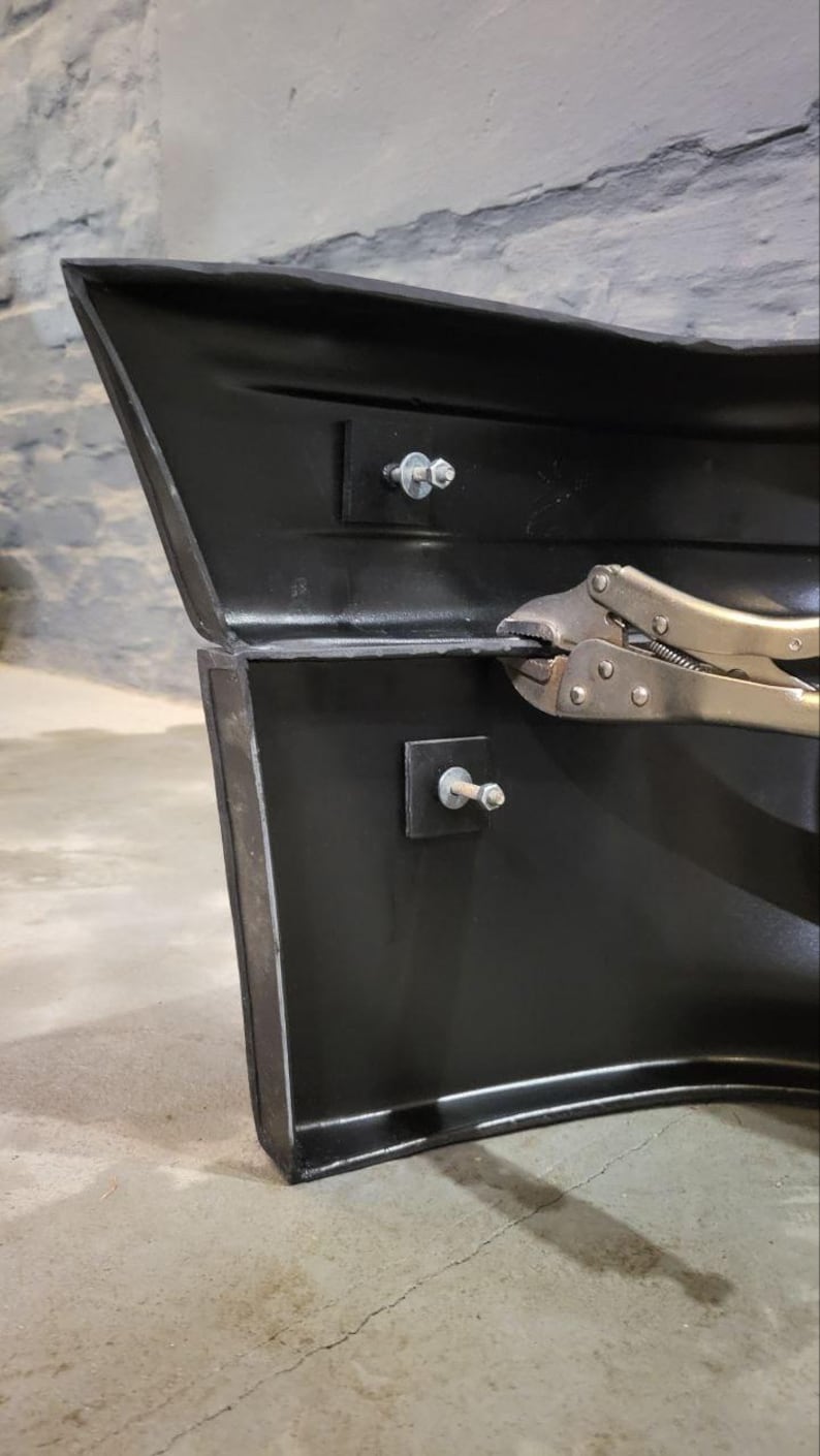 Puede incluir: Primer plano de una secci&oacute;n de parachoques automotriz negra con sujetadores met&aacute;licos. Un par de alicates est&aacute; sujeto al borde del parachoques. El parachoques est&aacute; contra una pared de ladrillos gris.