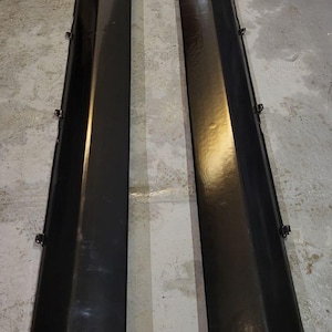 K&ouml;nnte beinhalten: Zwei schwarze, gl&auml;nzende Seitenschweller f&uuml;r Autos. Jeder Schweller ist lang und rechteckig mit einer leicht abgewinkelten Oberkante. Mehrere kleine Clips sind an den Seiten sichtbar. Die Teile sind wahrscheinlich f&uuml;r das &Auml;u&szlig;ere eines Fahrzeugs bestimmt.