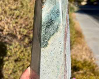May include: A green, white, and brown crystal point with a pointed top and a flat base. The crystal is polished and has a smooth surface.