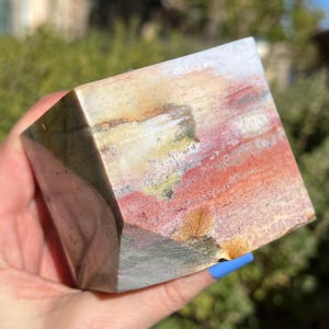 May include: A multi-colored, geometric stone cube with a polished surface. The cube displays a mix of red, white, brown, and green hues, with visible natural patterns. The stone is held in a hand, showcasing its size and shape.