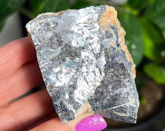 Può includere: Un grande cristallo naturale grezzo di colore argento, con una forma irregolare e ruvida. Il cristallo ha un'opalescenza metallica ed è parzialmente traslucido.