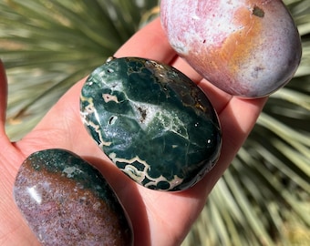 Può includere: Tre pietre preziose lucide nei toni del verde, viola e rosa sono tenute in una mano. Le pietre hanno forme lisce e arrotondate e motivi diversi. Lo sfondo è una vista sfocata di fogliame verde.