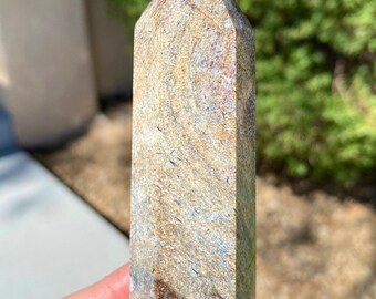 May include: A brown, gray, and white striped crystal point with a pointed tip. The crystal is about 4 inches long and is held in a hand.