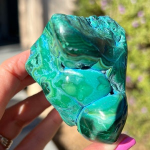 May include: A vibrant, raw malachite stone with a polished, glossy surface. The stone displays a rich, swirling pattern of deep emerald green and turquoise blue hues. The natural mineral formation is held in a hand, showcasing its unique texture and color variations.