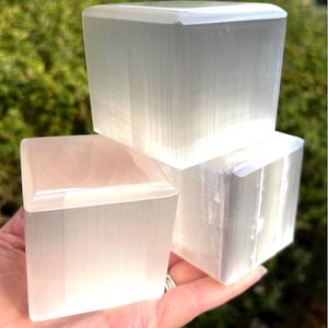 May include: Three white, translucent, cube-shaped selenite stones with a smooth, polished finish and visible vertical fibrous textures. The cubes are stacked and arranged together, displaying a bright, milky appearance with subtle light reflections.