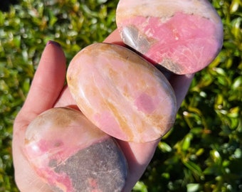 May include: Three pink, yellow, and gray polished stones with a smooth, oval shape. The stones are held in a hand.